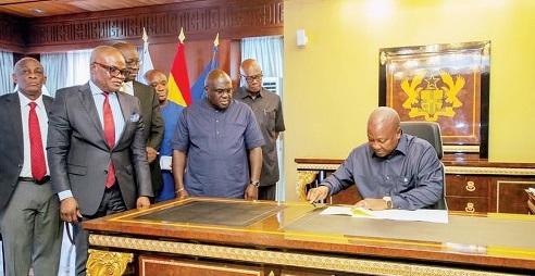 President John Mahama appending his signature to the Income Tax Amendment Act, while Julius Debrah (standing 2nd from right), the Chief of Staff, and other dignitaries look on
