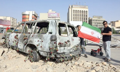 A man holding an Iranian flag by an Iranian Red Crescent ambulance that was destroyed during an Israeli strike