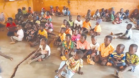   Pupils sitting on the bare floor with a visible crack in the classroom